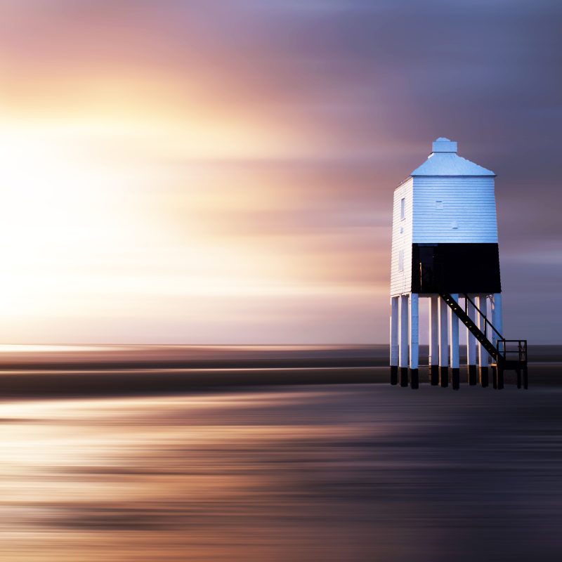 wooden lighthouse on stilts on a sandy beach with a glow of the setting sun behind