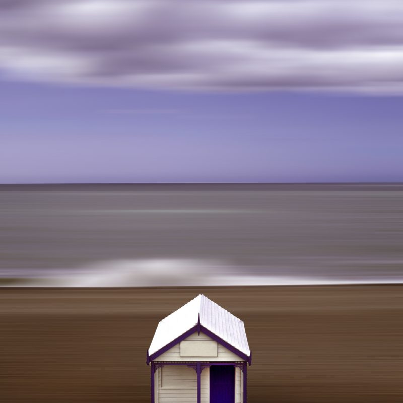 white beach hut on Brighton beach with wave crashing behind it.