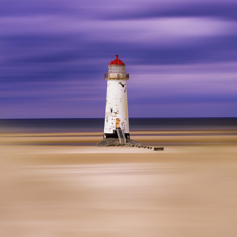 a white lighthouse with a red top on a sandy beach with a blue sea and sky behind