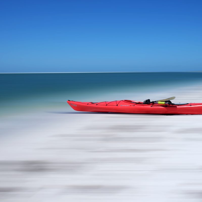 A red Kayak on a white sandy beach with blue sea and sky behind