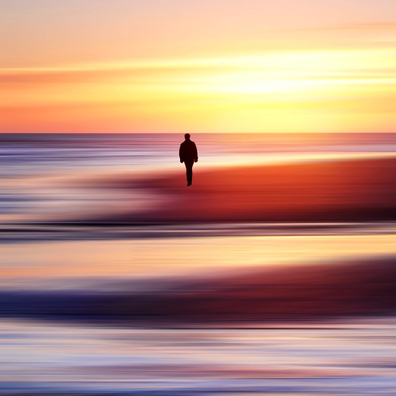 solitary person silhouetted walking along sandy beach at sunset with the sea in the background