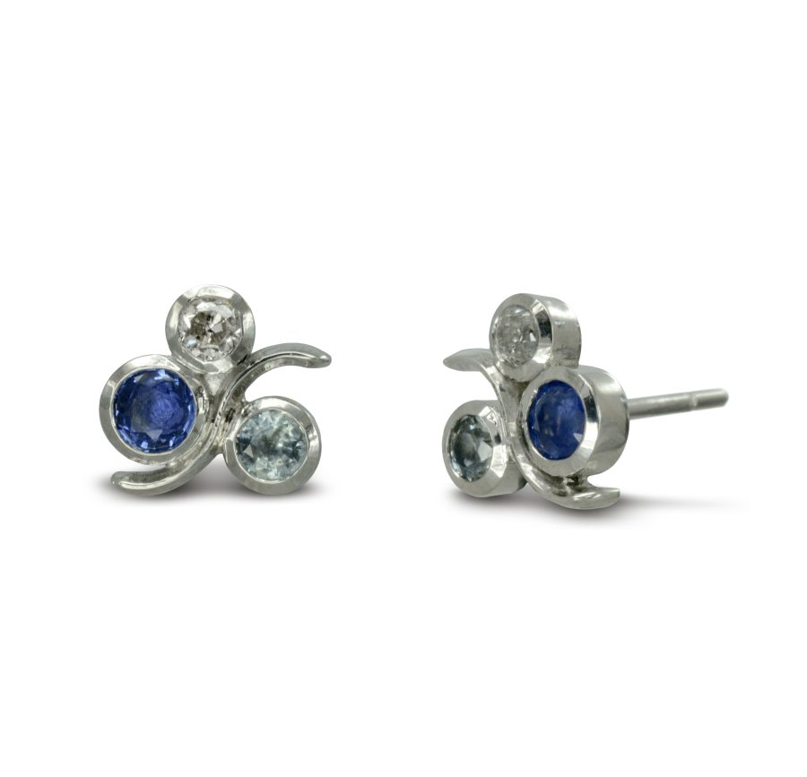 A platinum, sapphire and diamond ear studs: two round blue sapphires in different sizes and shades, with a diamond set in swirling platinum metalwork. Inspired by water and bubbles.