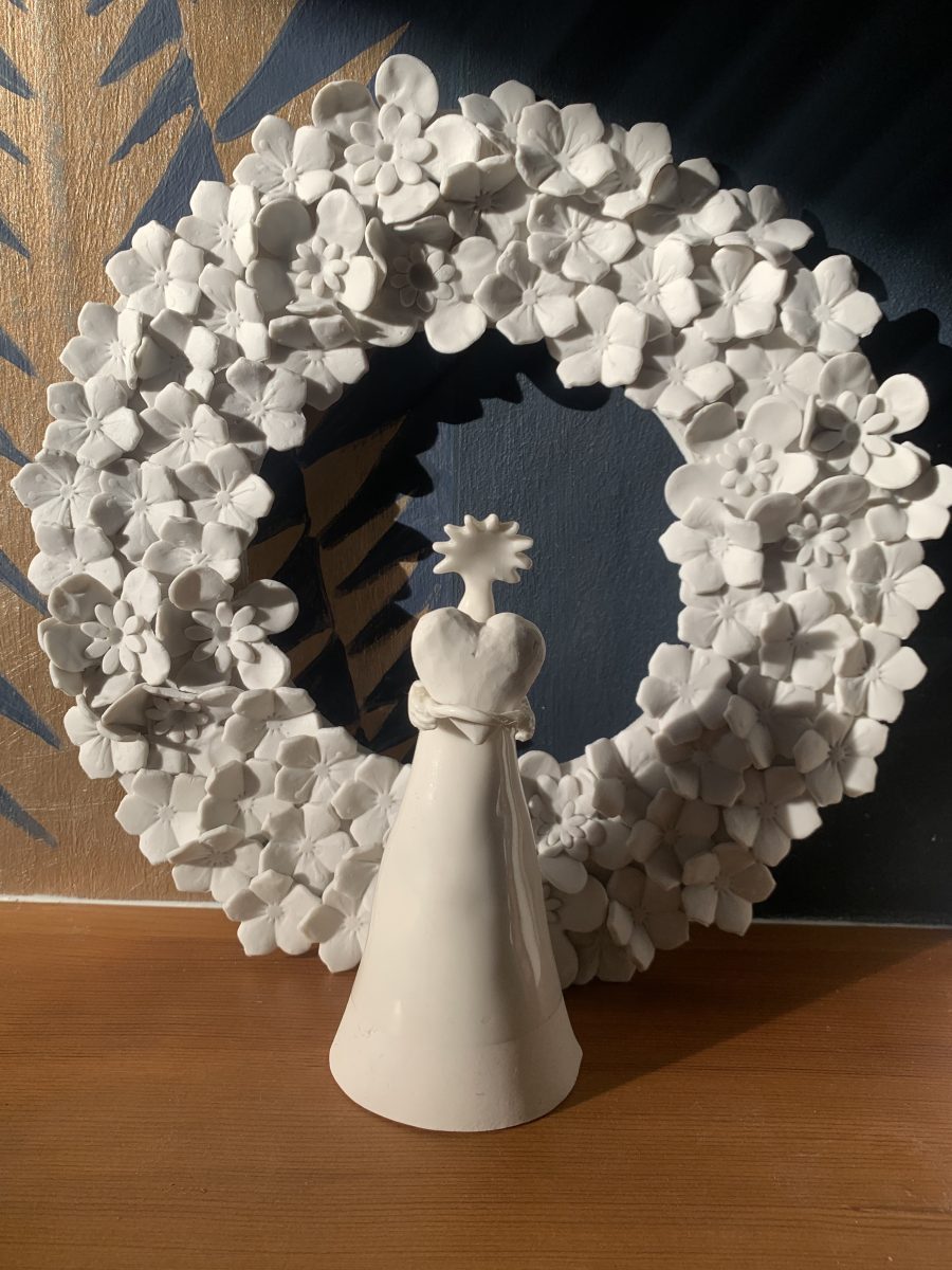 Large white porcelain wreath with a porcelain  Angel holding a large heart in her hands standing in front of the wreath . All placed on a fireplace mantelpiece. 
