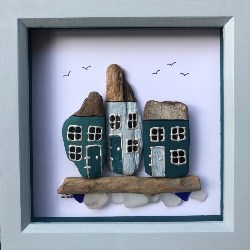 3 driftwood houses painted in shades of blue with a seaglass ‘sea’ below them. Framed in a deep box frame painted in coordinating colours.