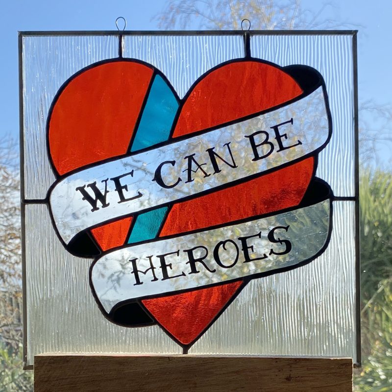 A stained glass ornament of a heart with a scroll saying 'we can be heroes' across it