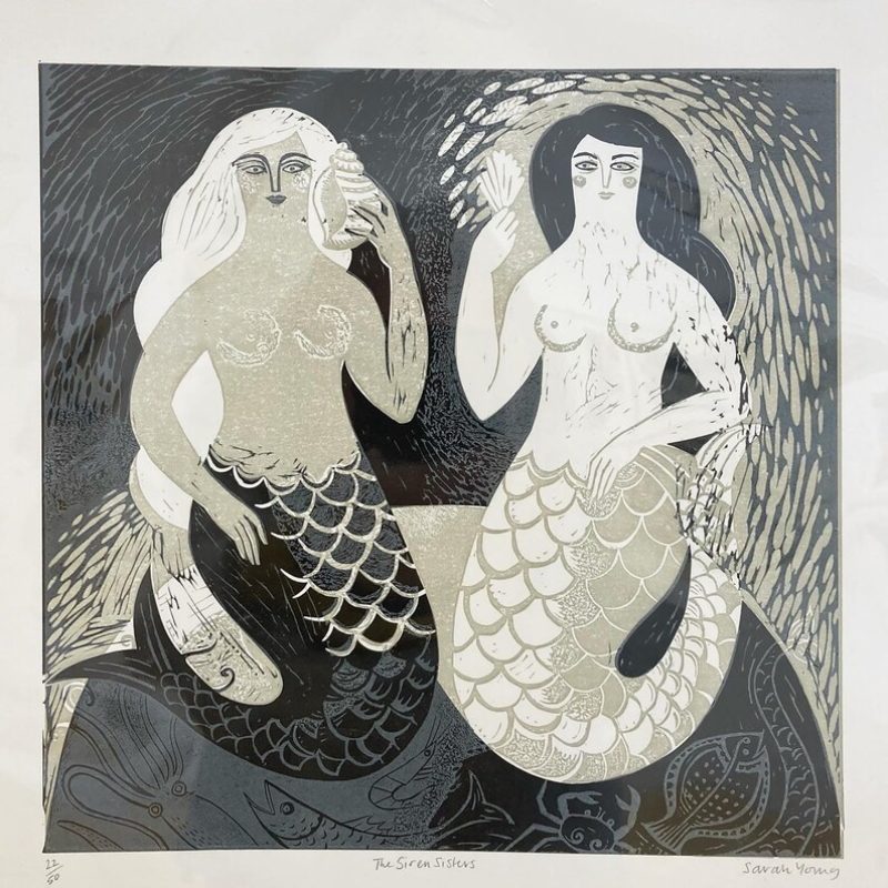 A relief print of two mermaids sitting on a rock together in the sea
