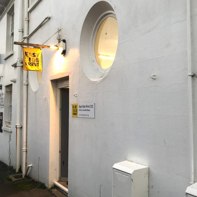 The outside of the East Side Print studio, with our yellow flag hanging above the door. There are two round windows above the door to the studio.