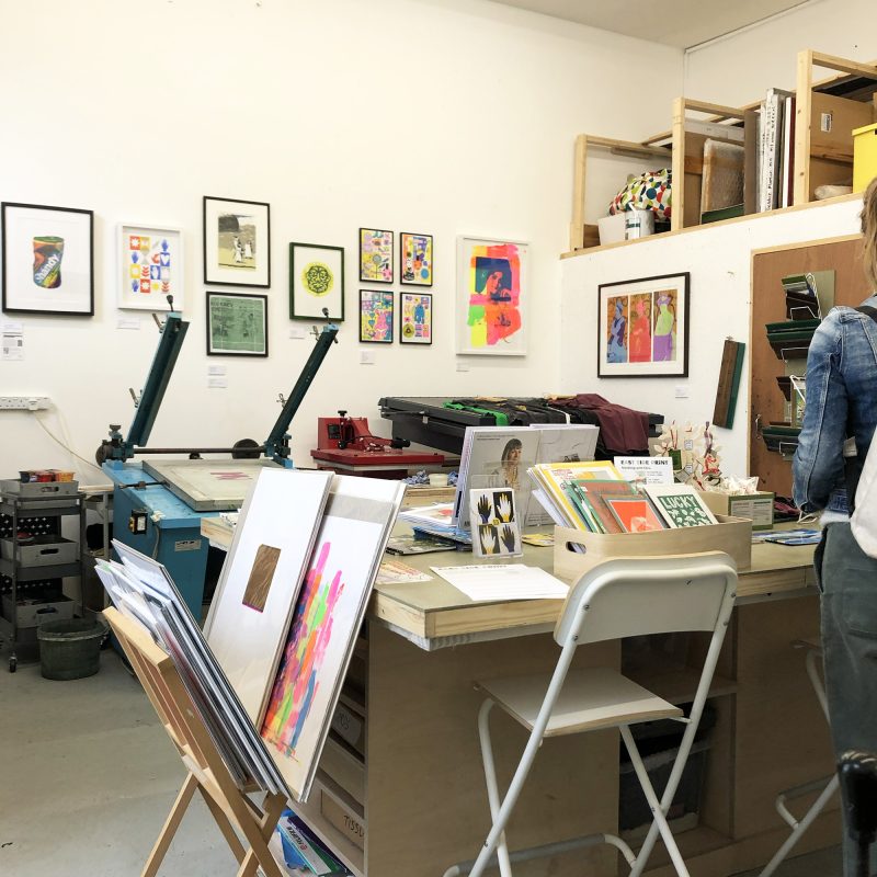 The East Side Print studio set up for AOH 2022, with framed prints on the wall and printed items like tea towels, cards and t-shirts laid out on a table.