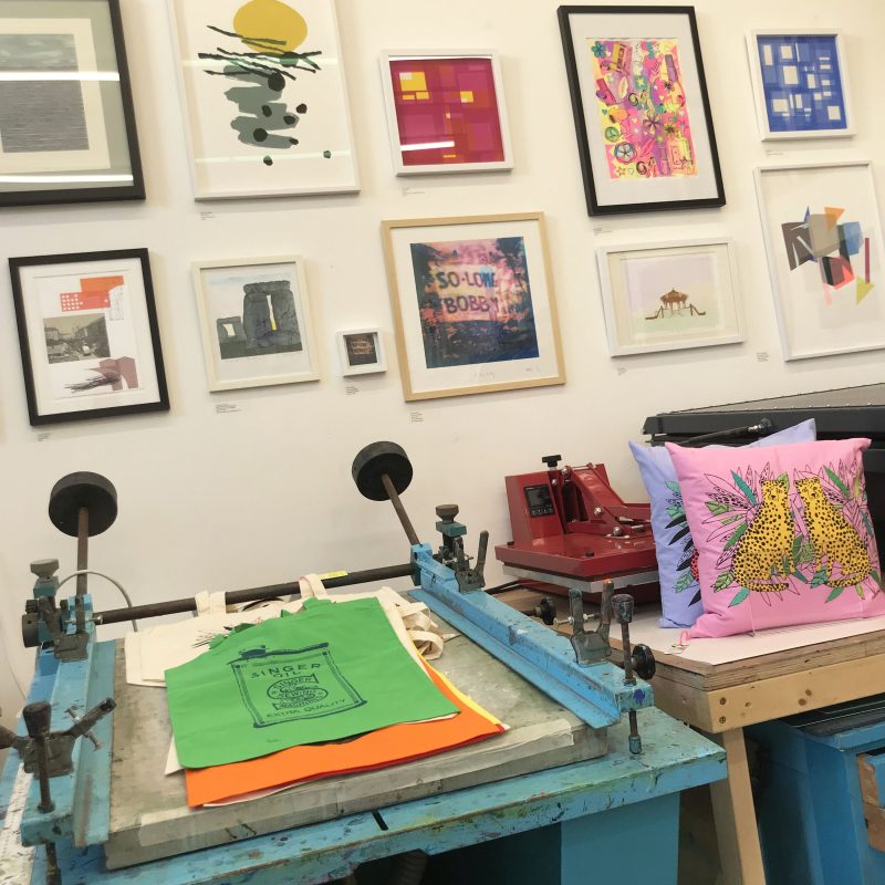 Framed prints hung on the wall in the East Side Print studio, with printed tote bags laid out on a screenprinting bed in the foreground. 