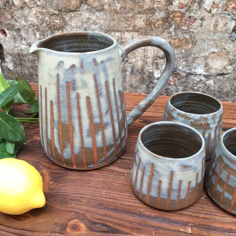 A selection of contemporary rustic ceramic vessels on a wooden board. These include a jug and 2 mugs.