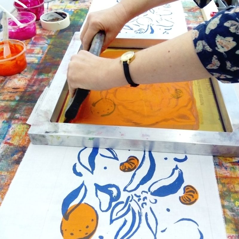 A length of fabric is laid out on a table and someone is screenprinting onto it with orange ink.