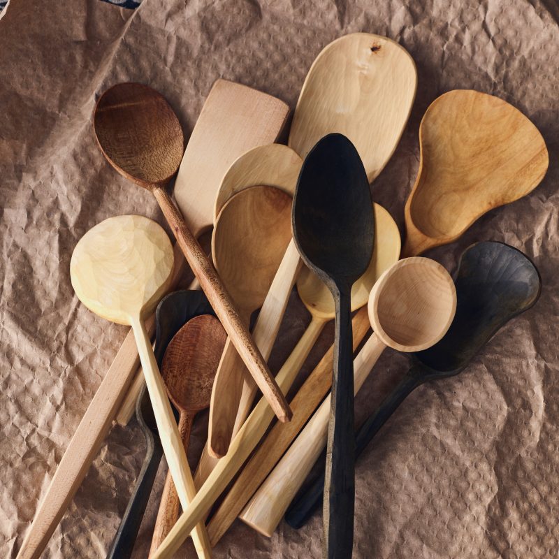 a collection of wooden spoons hand carved by Annie slack. the wood colours range form a palest cream right through to dark blackish brown. they lie on top some brown packing paper.