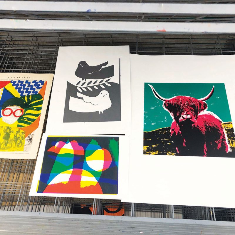 4 screenprints are drying on a rack in the East Side Print studio. One is a grey print of a bird, one shows a highland cow printed in the style of Andy Warhol, the third is an abstract image made by overprinting cyan, magenta and yellow ink. The final print is an image of a face surrounded by pattern, printed on a page of a book in red, yellow and blue inks.