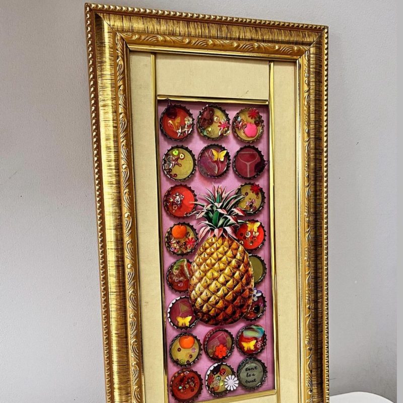 Vintage gold box frame with pink background, bottle caps filled with pink, Orange and yellow recycled materials and resin, and a collage pineapple in the middle. 