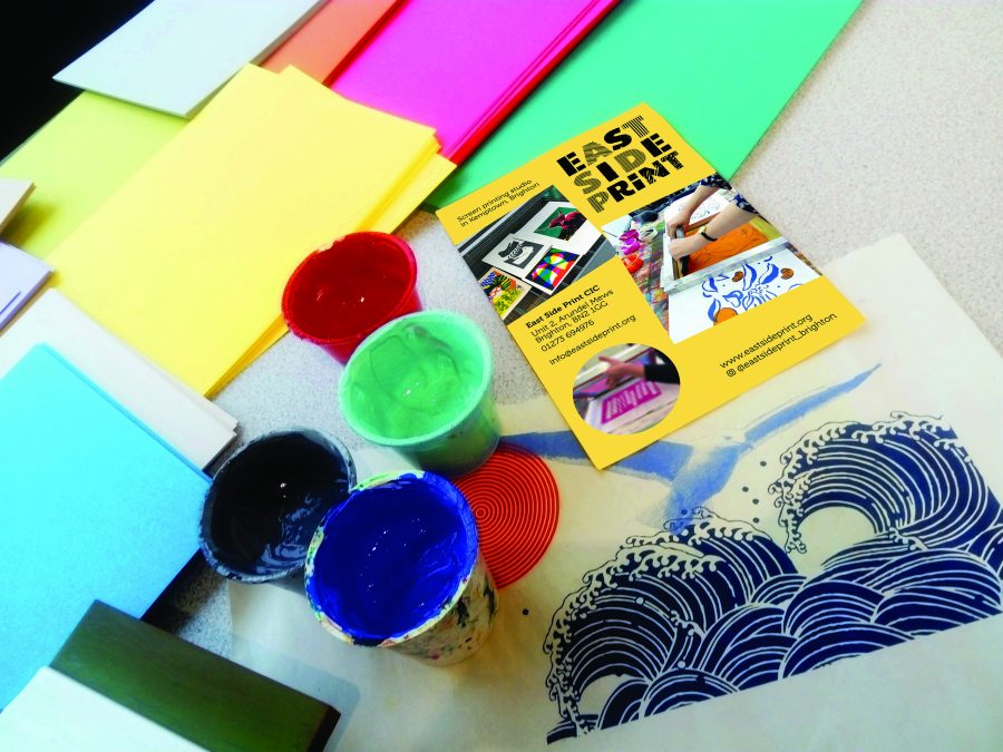 A table top strewn with pots of screenprinting inks and coloured paper. In the foreground is a screenprint of a blue wave, and above this is an East Side Print flyer.