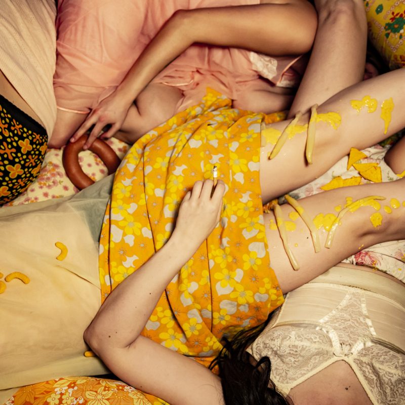 A photo of bodies lying together, dressed in clothes with a 70’s style and Hugh, a hand lets cigarette ash drop onto spaghetti hoop stained clothes.