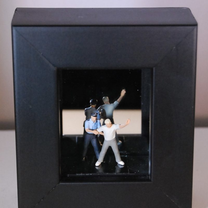 a 1/87 scale scene of a man being arrested by a policeman,in a small wooden box frame with a mirrored back