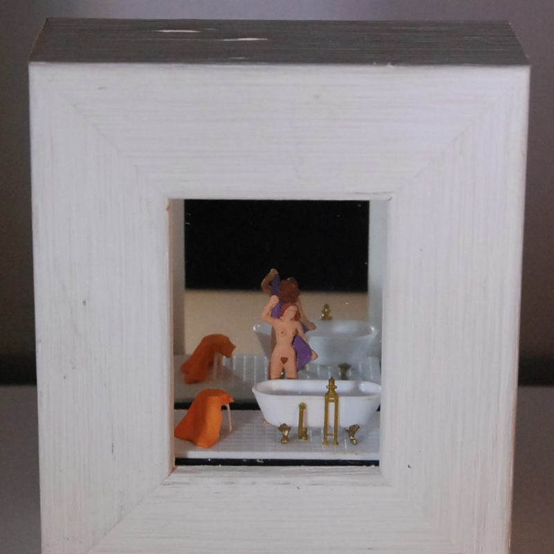 a 1/87 scale scene of a naked woman facing  forwards standing by an old fashioned bath towelling herself dry in a small white wood box frame with a mirrored back. The piece is about  6 x 4 inches