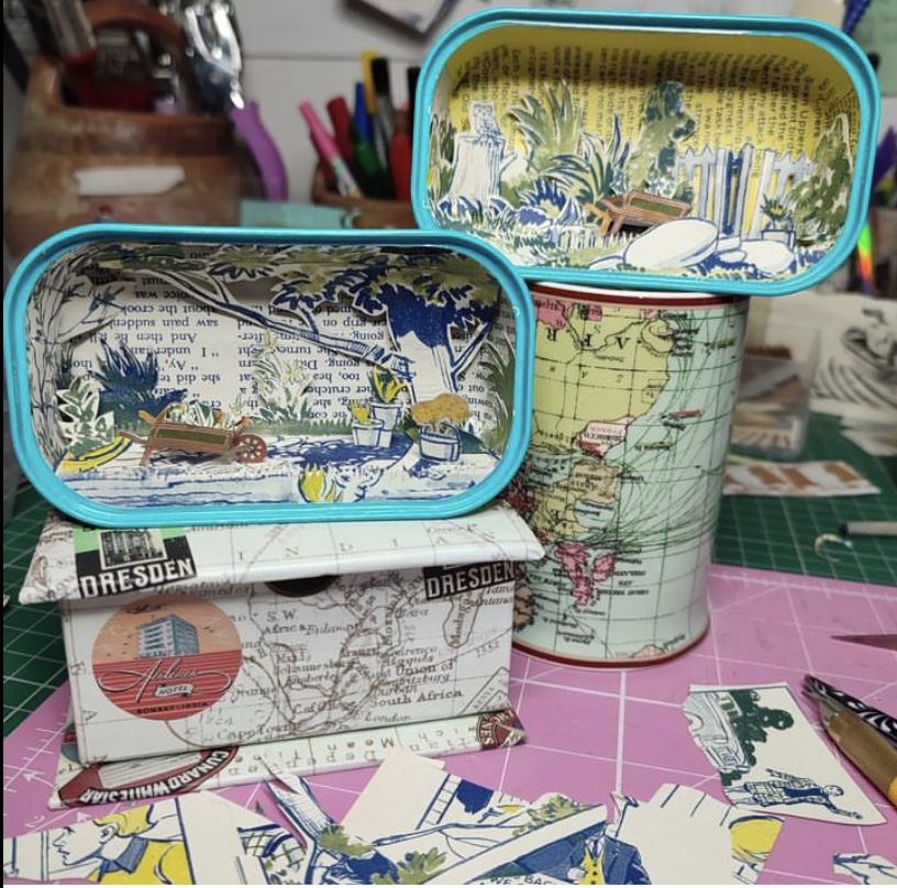 Sardine cans with a diorama of an English country garden scene made of paper.