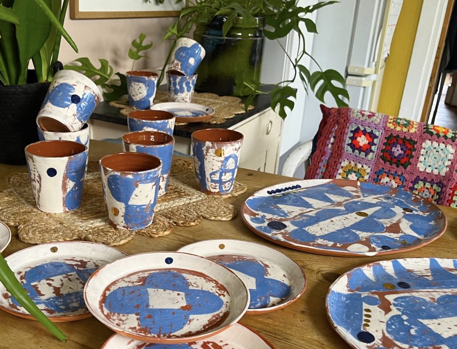 in this photo you can see a collection of blue and white boldly patterned tumblers, plates and platters arranged on a wooden table. in the back ground is a leafy houseplant and a chair with a brightly coloured crocheted blanket.