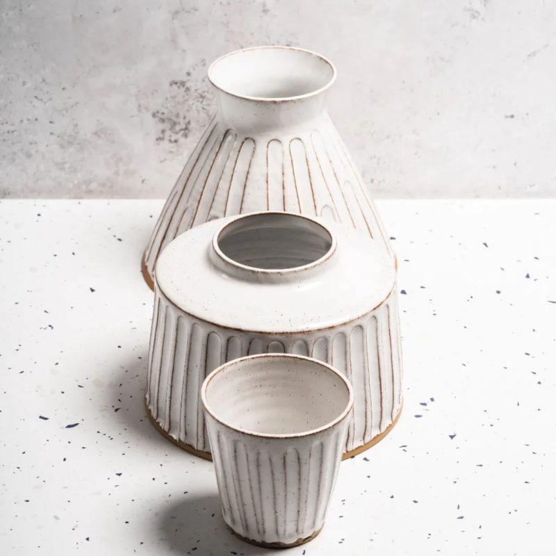 3 white glazed pots lined up in front of each other. in front is a small tumbler and the behind are two vases one slightly smaller and with a short neck and the one furthest behind taller with a funnel neck. they all are decorated with vertical grooves.