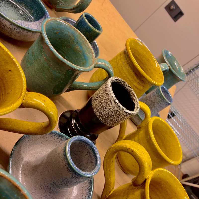 Multiple mugs and both curved/straight candle holders are playfully arranged, creating a colourful scene full of mustard yellow, speckled silver and turqoise blue cover pots.