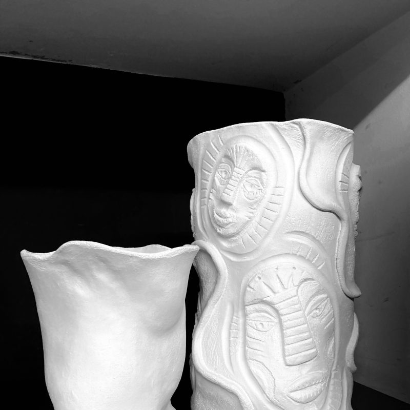 Two vases stand side by side, in a black and white image. The right tall and slender, decorated with multiple engraved faces and a single relief line snaking its way around the forms. The left shorter with the surface left bare and a slight floral appearance.