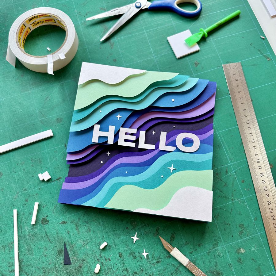 A photograph of a papercut layered artwork, with celestial waves, stars and the word 'HELLO'. In the background is a cutting mat, scalpel and other tools