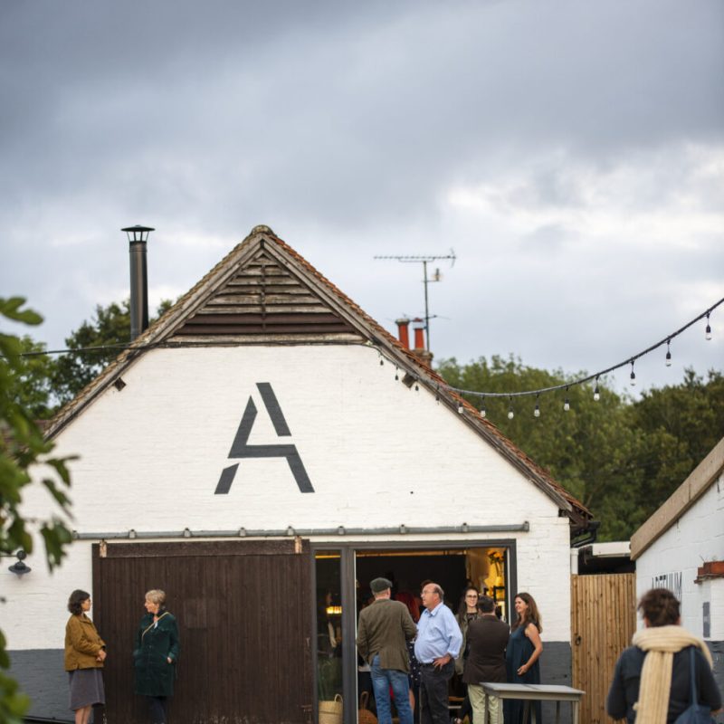 A white converted barn with an A painted on it. People are milling outside and spilling out of the doors. 
