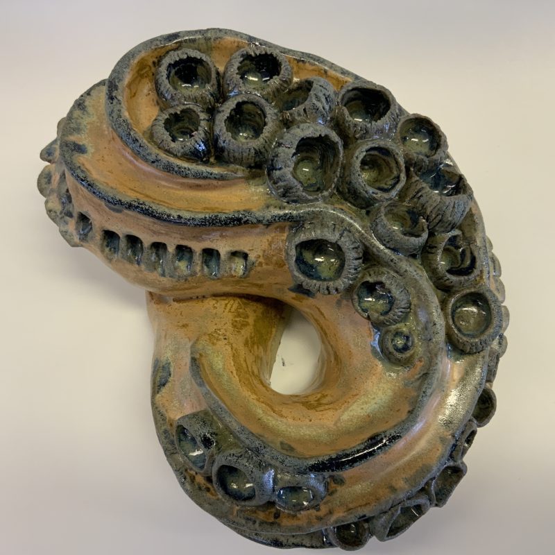 A ceramic piece in a figure of 8 shape in blues. greens and yellows with circular pieces attached at one end.