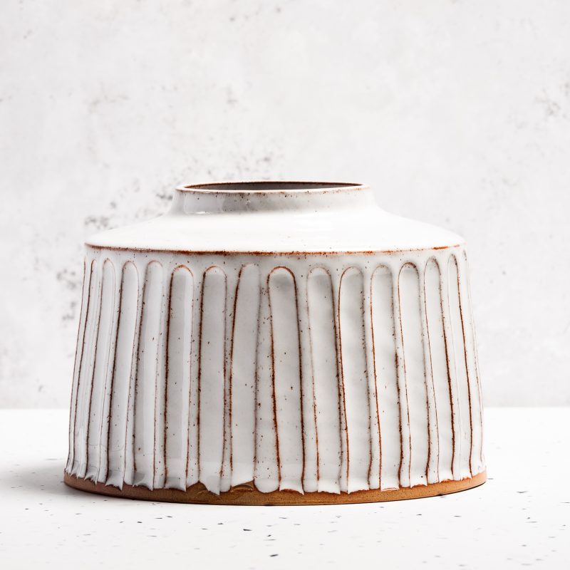 A stoneware vase finished in a glossy white glaze with textured vertical lines. The vase measures approximately 14 cm in height and 18 cm in diameter and looks like an old-fashioned flaggon in shape