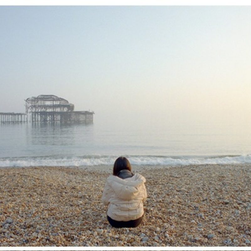 Loan girl in cream anorak sits on pebbled beach., before the West Pier. 