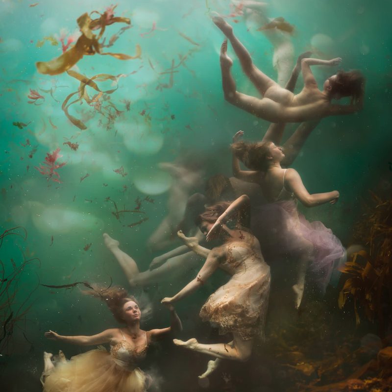 A photograph taken underwater, showing a small group of women. They are dancing, floating and rising in the turquoise sea, with pieces of seaweed surrounding them.  The image has a baroque feel and is inspired by heavenly scenes in 18th century ceiling frescos.