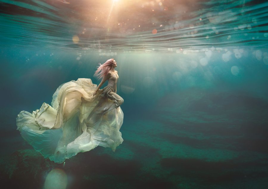 A photograph of a deep blue underwater scene taken near the island of Formentera in the Mediterranean Sea. In the image there is a young woman who is gracefully floating a few metres deep under the surface. She has long pale pink hair and she is wearing a voluminous silk parachute skirt which is flowing behind her. The sun is shining through the surface of the water and creating striking beams of light.