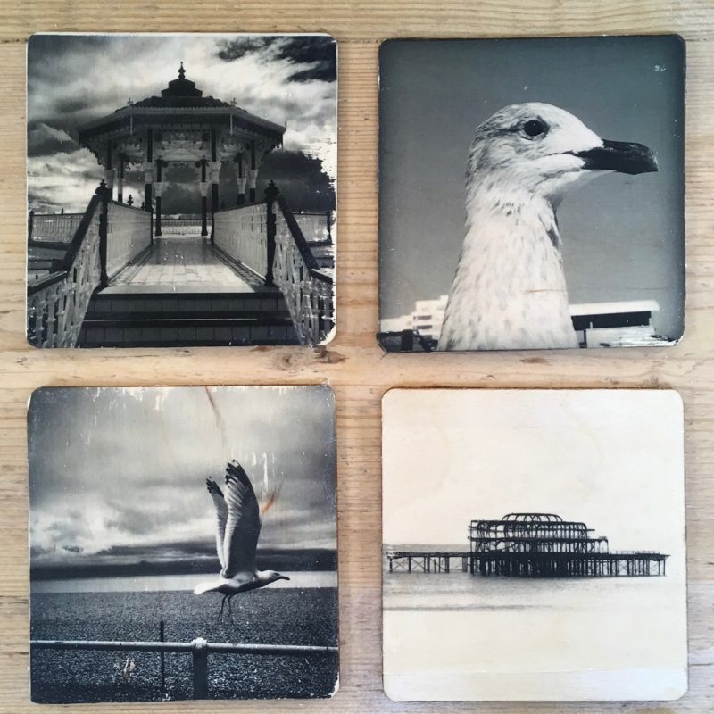 Four original black and white photographic images, transfer printed by hand onto wood. Images are of Brighton Bandstand with dramatic clouds behind; close up profile of a seagull; seagull taking off from railing against backdrop of the beach, sea and ‘moody’ sky; high contrast shot of West Pier from a side angle.