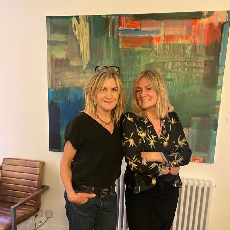 Two women stand close together - a large green painting is behind them