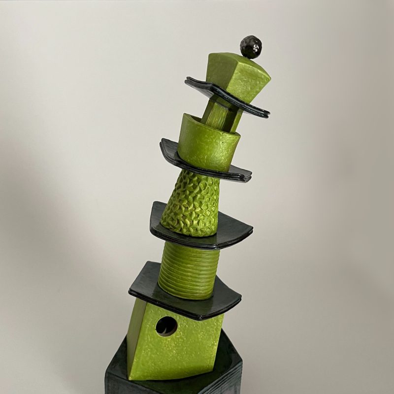 A sculpture made of ceramic in the form of a teetering stack of washing up or a leaning pottery pagoda. Each of the five lime green sections is separated by a shiny black pentagonal plate or shallow dish like a saucer.