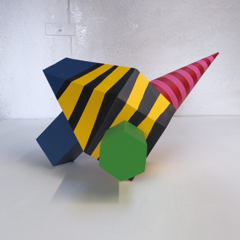 A painted wooden sculpture with a central house shaped structure painted with black and yellow stripes. Pink and red cone is protruding on the right at the back, a green hexagonal box is stuck on the front towards the right and a dark box form supports the rear at the left.