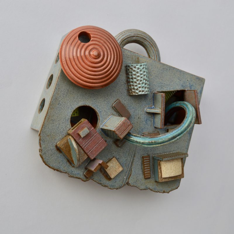 A ceramic wall sculpture in the form of a suitcase with handle like accretions and maybe a small city attached
