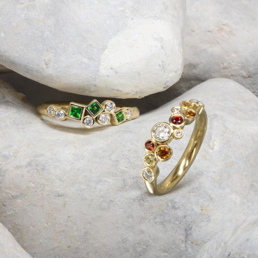 Two gold half eternity rings, one with round diamonds and square emeralds in a long cluster formation, and one with round fire sapphires and diamonds in a long cluster formation.