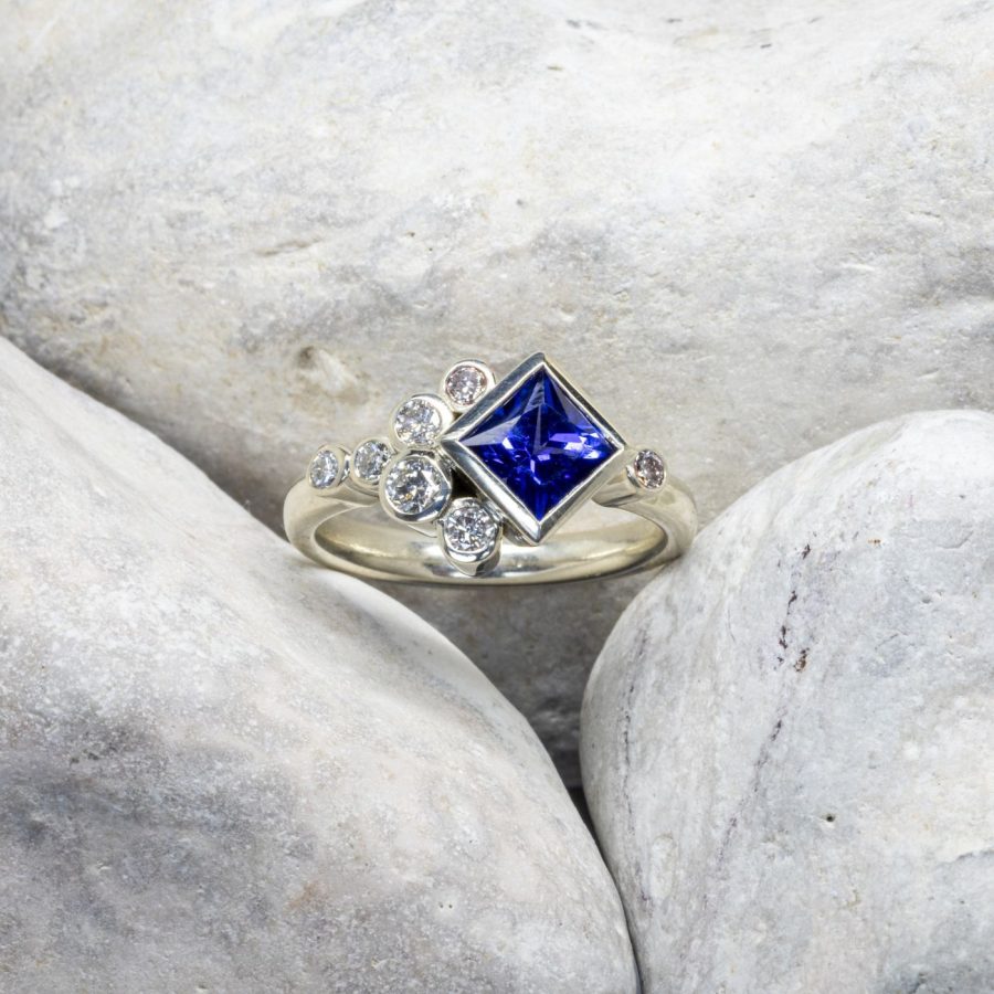 A princess cut Tanzanite and smaller round diamonds in rubover settings clustered together, all set in Platinum