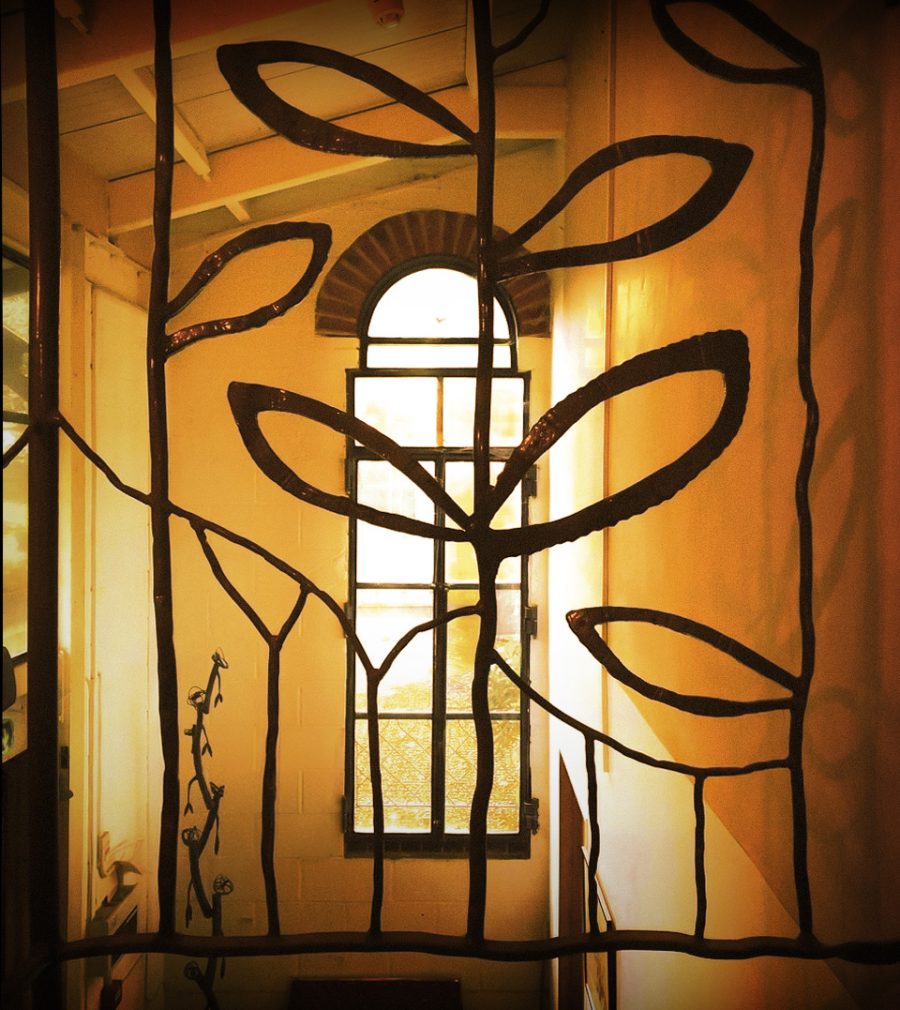 A view through a decorative metal balustrade onto the hallway of the main building with glistening light coming through a tall window. 