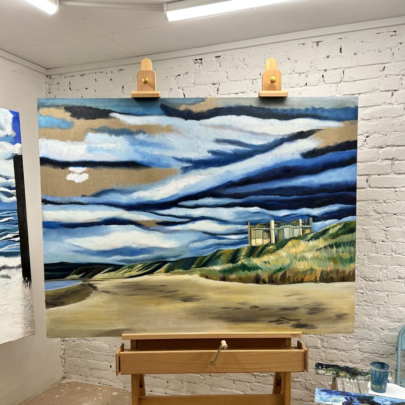 A work in progress: A large beach painting with cloudy sky and castle-like building. 