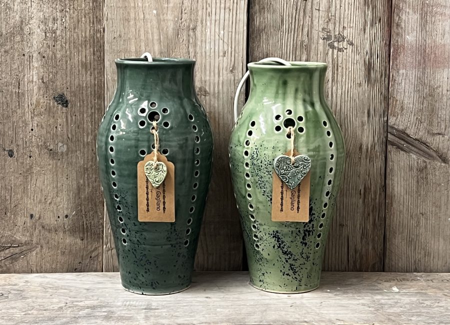 The image shows two handcrafted pottery lamps, both made on the pottery wheel. The lamps have small, carefully placed holes throughout, allowing light to shine through when lit. They are glazed in two distinct shades of green—one a deep forest green, and the other a lighter, earthy tone. The combination of the smooth, rounded shape and the green glaze creates a natural, calming aesthetic.