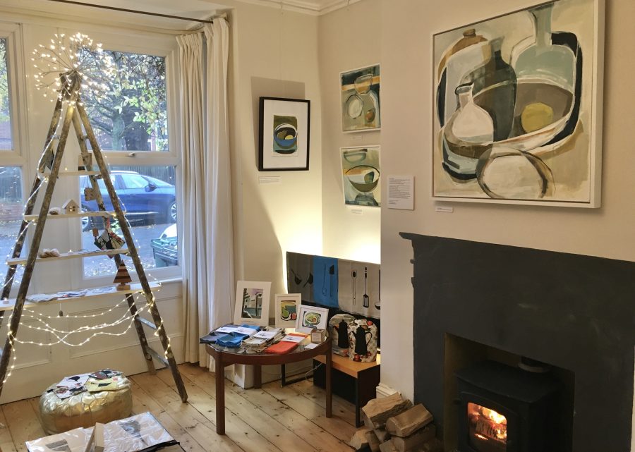 An interior living room space with a lit fireplace, decorative ladder shelf with fairy lights, paintings on the walls and textiles displayed on a table.