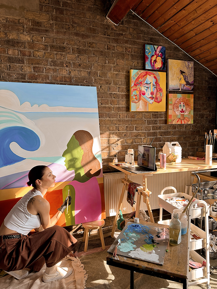 Rocio in her studio painting a large ocean themed work with various paintings on an exposed brick wall.