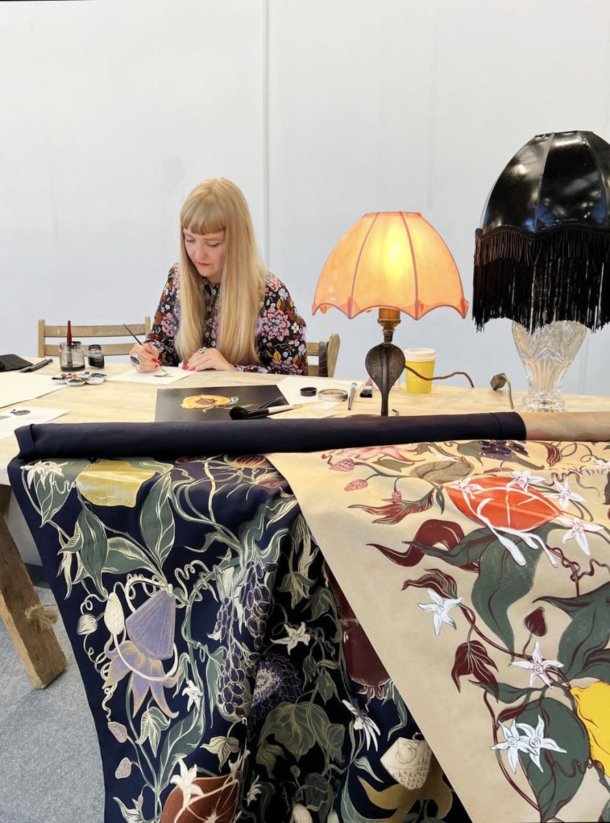 A photograph of Rosanna Bishop painting, alongside fabrics screen-printed with fruits and latex lampshades.