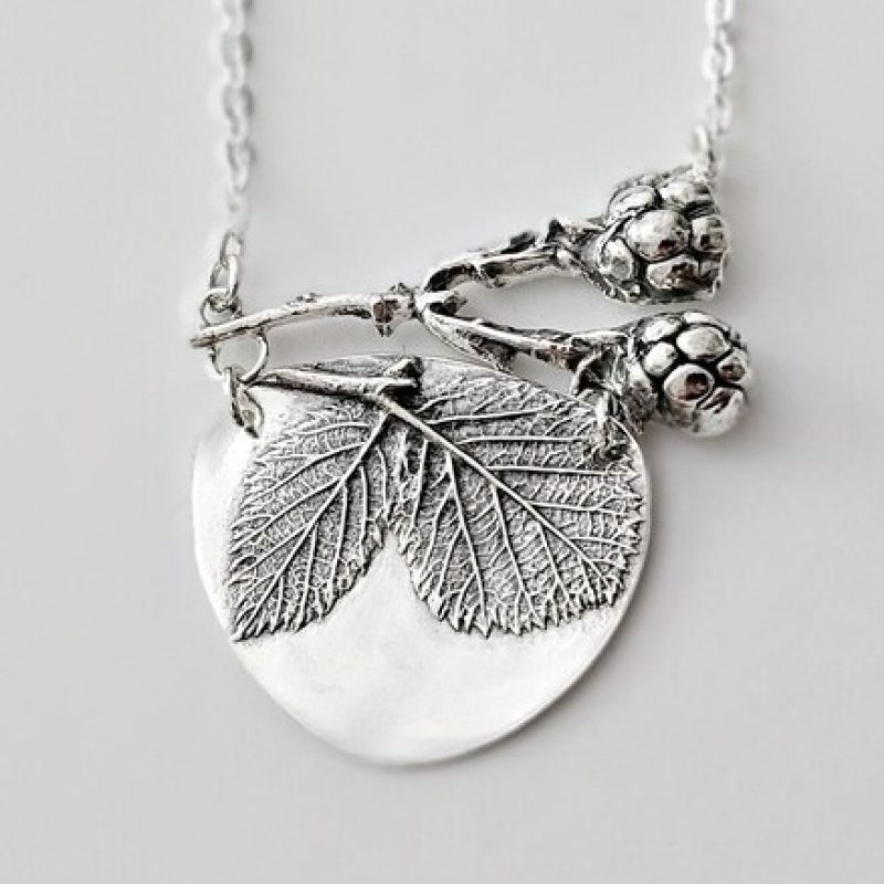 The image features a silver necklace with a nature-inspired pendant. The pendant includes two detailed leaves, a small branch, and textured blackberry fruits. Attached to a delicate silver chain, the design has a vintage, botanical aesthetic, highlighting nature's beauty.