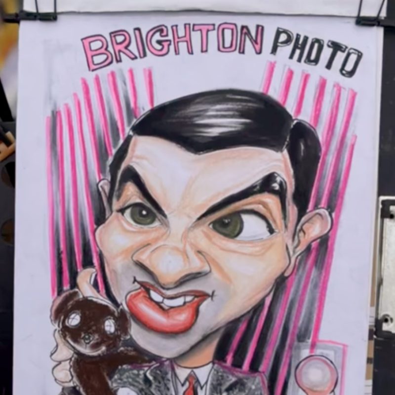 Describe the joyful moment image of Mr Bean taking his selfies on the photo booth.. This is a hand-drawn caricature of Mr Bean with exaggerated features, a goofy grin and expressive eyes. He holds his teddy bear in one hand and a strip of photos in the other, set against a pink lined background. The words 'BRIGHTON PHOTO' appear above, suggesting a fun coastal setting. The artwork uses mixed media with bold colours and rich textures, bringing his quirky personality to life.