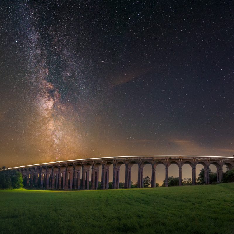 The Milky Way core towers above the Balcombe Viaduct as the lights from a train heading to London pass across the bridge. A small meteor can be seen just to the right of the Milky Way.
